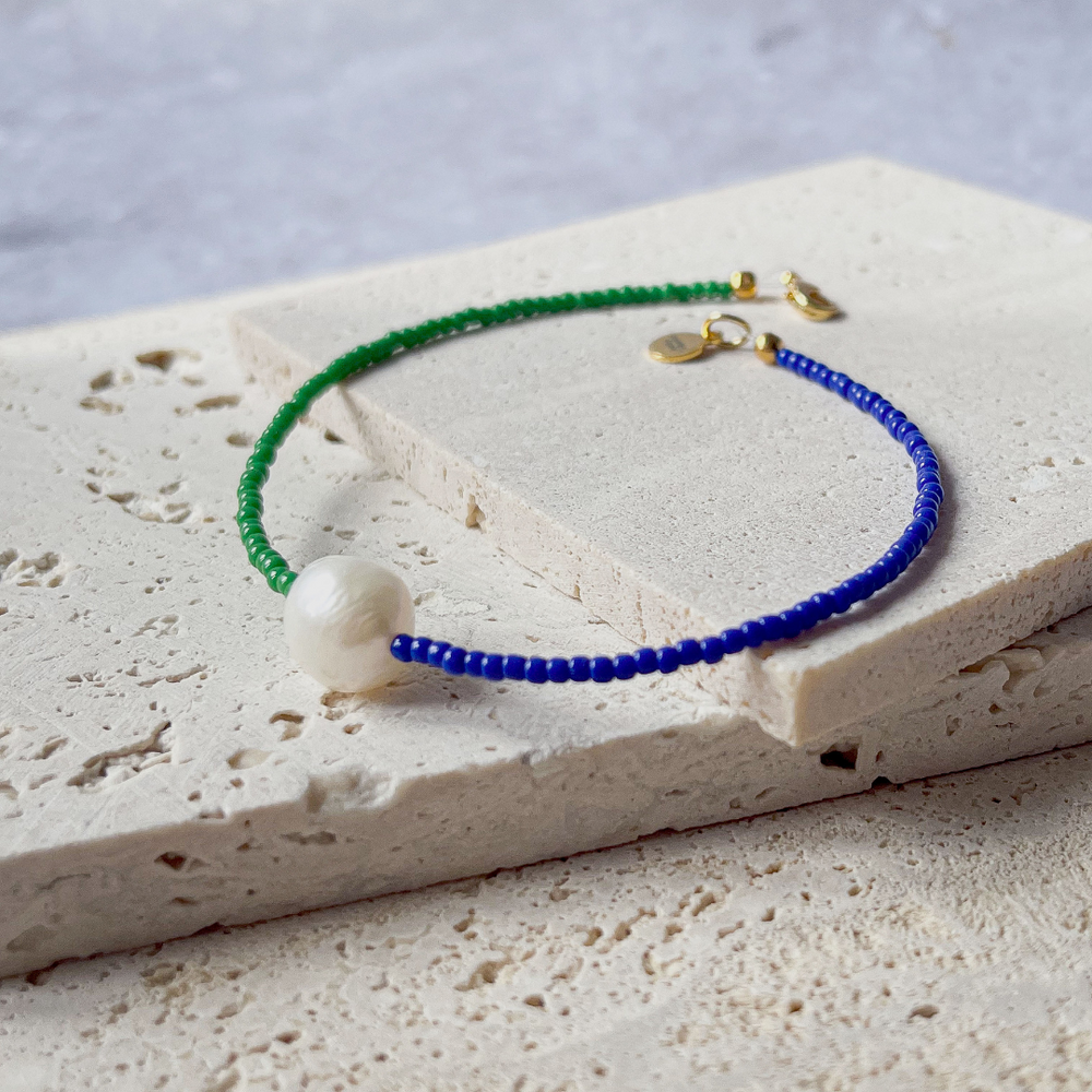 A minimalist bracelet featuring a large single baroque pearl on a string of small midnight blue and forest green glass beads, presented on a corner of two textured aerated concrete blocks. The blocks are placed against a grey background, highlighting the necklace's delicate colors and elegant design. The large pearl serves as a focal point, giving the piece a touch of classic sophistication mixed with a hint of modern color contrast.