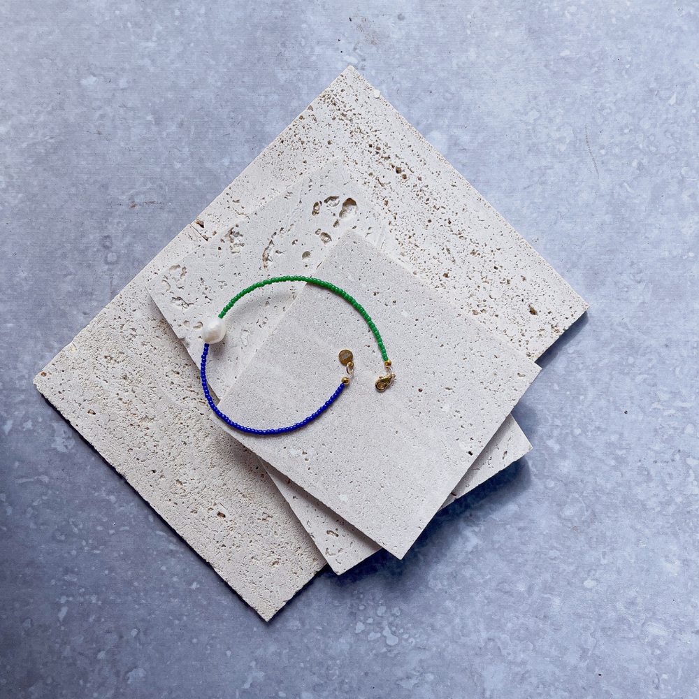 A minimalist bracelet featuring a large single baroque pearl on a string of small midnight blue and forest green glass beads, presented on a corner of two textured aerated concrete blocks. The blocks are placed against a grey background, highlighting the necklace's delicate colors and elegant design. The large pearl serves as a focal point, giving the piece a touch of classic sophistication mixed with a hint of modern color contrast.