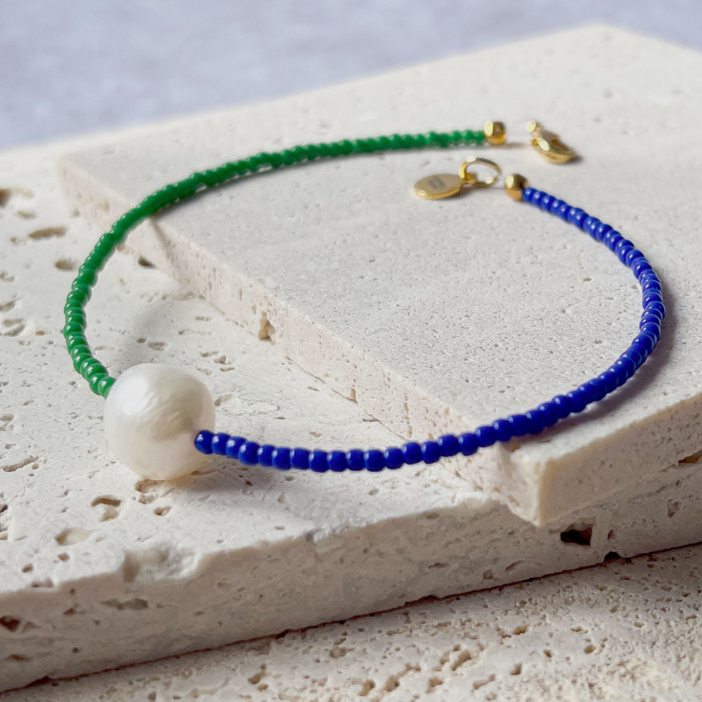 A minimalist bracelet featuring a large single baroque pearl on a string of small midnight blue and forest green glass beads, presented on a corner of two textured aerated concrete blocks. The blocks are placed against a grey background, highlighting the necklace's delicate colors and elegant design. The large pearl serves as a focal point, giving the piece a touch of classic sophistication mixed with a hint of modern color contrast.