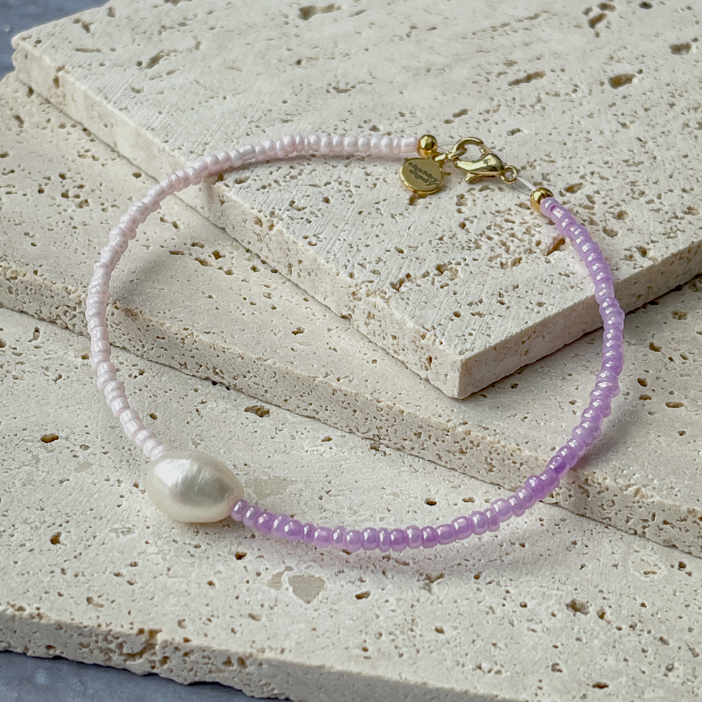 A minimalist bracelet featuring a large single baroque pearl on a string of small white and lavender glass beads, presented on a corner of two textured aerated concrete blocks. The blocks are placed against a grey background, highlighting the necklace's delicate colors and elegant design. The large pearl serves as a focal point, giving the piece a touch of classic sophistication mixed with a hint of modern color contrast.