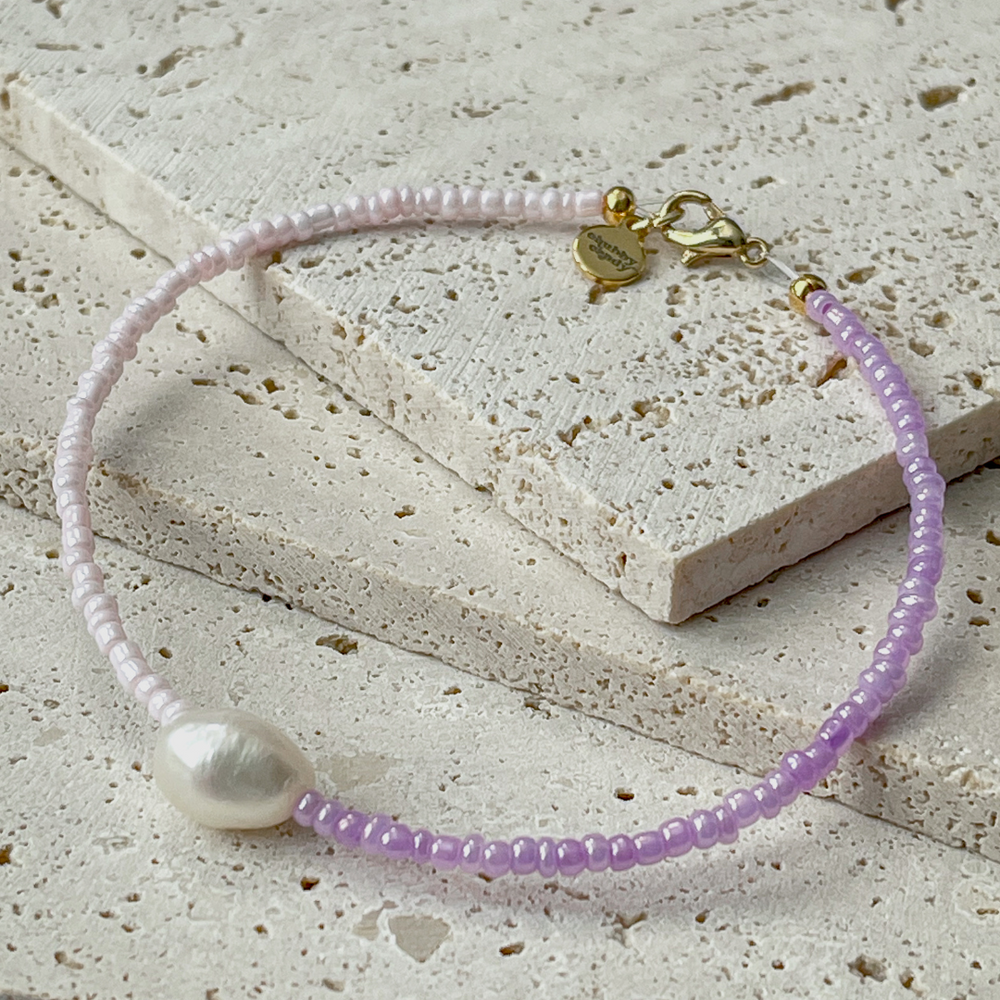 A minimalist bracelet featuring a large single baroque pearl on a string of small white and lavender glass beads, presented on a corner of two textured aerated concrete blocks. The blocks are placed against a grey background, highlighting the necklace's delicate colors and elegant design. The large pearl serves as a focal point, giving the piece a touch of classic sophistication mixed with a hint of modern color contrast.