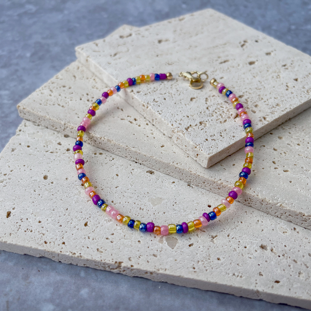 A vibrant beaded bracelet with a rainbow of colors, secured with a gold-tone clasp, displayed on a light concrete block backdrop.