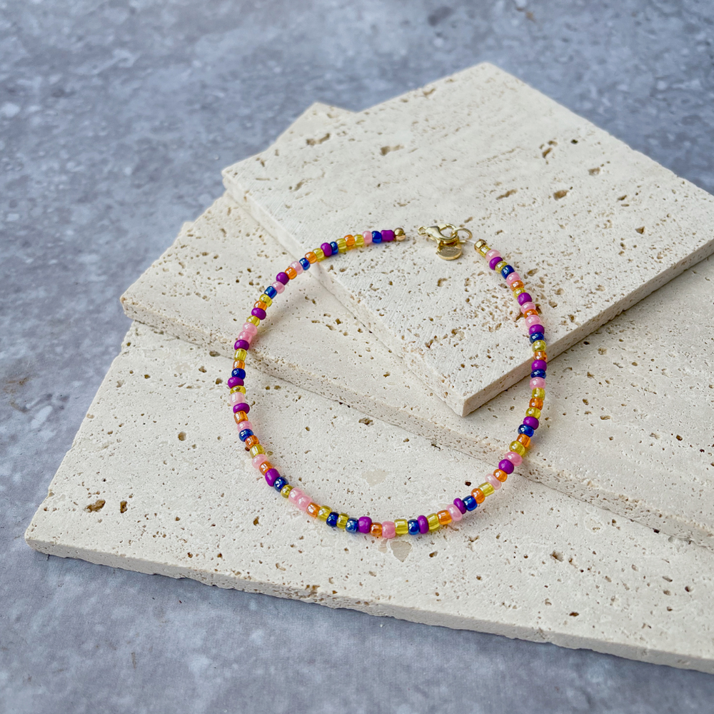 A vibrant beaded bracelet with a rainbow of colors, secured with a gold-tone clasp, displayed on a light concrete block backdrop.