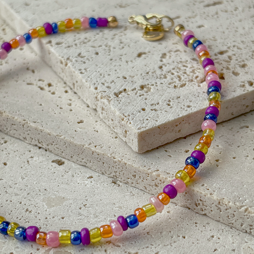 A vibrant beaded bracelet with a rainbow of colors, secured with a gold-tone clasp, displayed on a light concrete block backdrop.