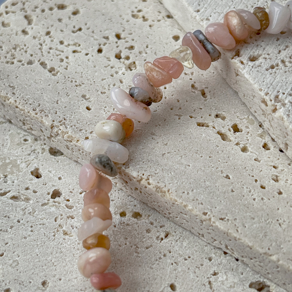 A bracelet featuring pink opal chips and bronze beads on aerated concrete. The opal chips vary in pink, white, and grey, complemented by the shine of bronze beads. It's fastened with a gold-tone clasp and brand tag, set against the concrete's neutral texture, emphasizing the bracelet's elegance.