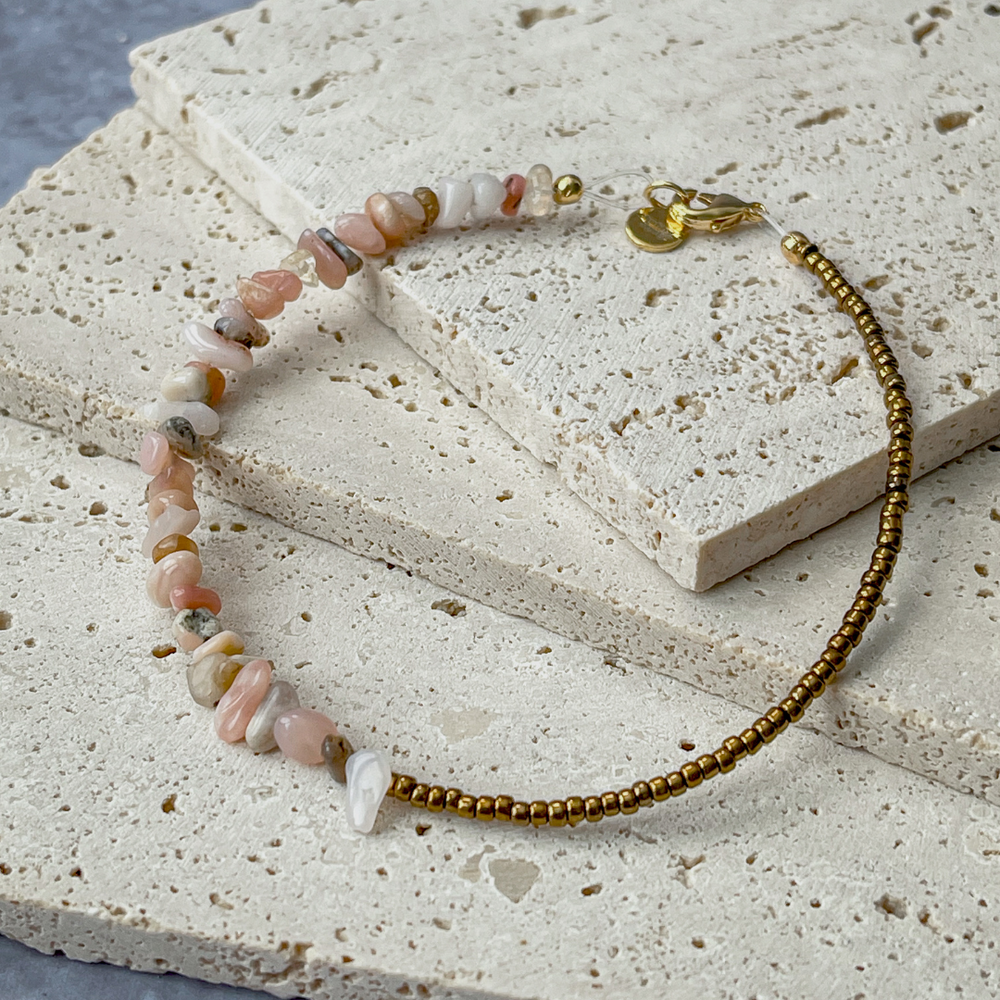 A bracelet featuring pink opal chips and bronze beads on aerated concrete. The opal chips vary in pink, white, and grey, complemented by the shine of bronze beads. It's fastened with a gold-tone clasp and brand tag, set against the concrete's neutral texture, emphasizing the bracelet's elegance.