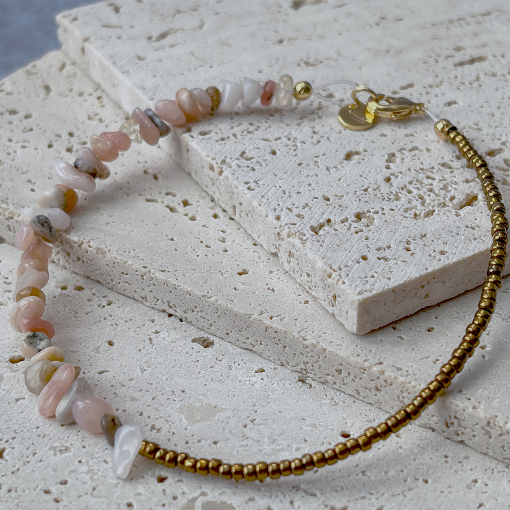 A bracelet featuring pink opal chips and bronze beads on aerated concrete. The opal chips vary in pink, white, and grey, complemented by the shine of bronze beads. It's fastened with a gold-tone clasp and brand tag, set against the concrete's neutral texture, emphasizing the bracelet's elegance.