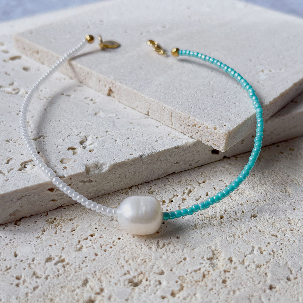 A minimalist bracelet featuring a large single baroque pearl on a string of small white and turquoise glass beads, presented on a corner of two textured aerated concrete blocks. The blocks are placed against a grey background, highlighting the necklace's delicate colors and elegant design. The large pearl serves as a focal point, giving the piece a touch of classic sophistication mixed with a hint of modern color contrast.