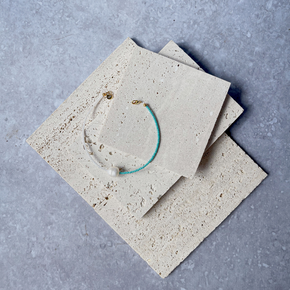 A minimalist bracelet featuring a large single baroque pearl on a string of small white and turquoise glass beads, presented on a corner of two textured aerated concrete blocks. The blocks are placed against a grey background, highlighting the necklace's delicate colors and elegant design. The large pearl serves as a focal point, giving the piece a touch of classic sophistication mixed with a hint of modern color contrast.