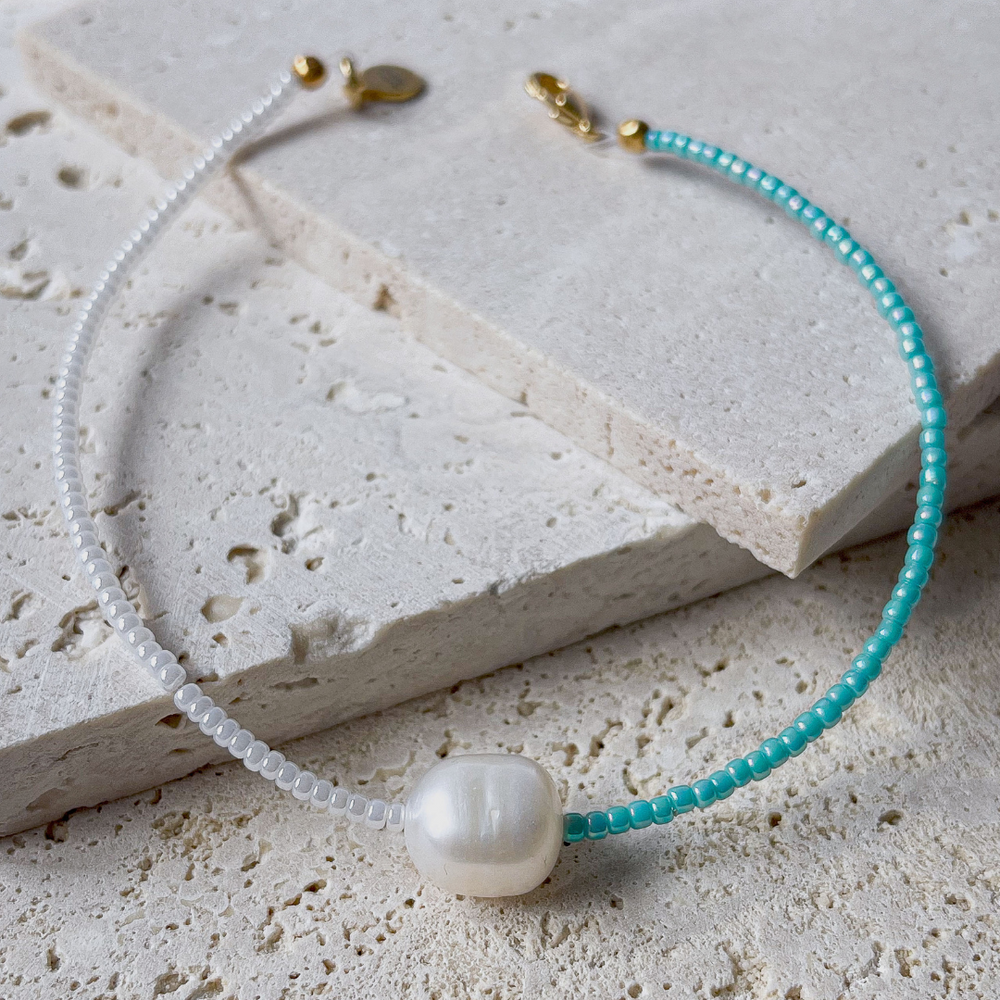 A minimalist bracelet featuring a large single baroque pearl on a string of small white and turquoise glass beads, presented on a corner of two textured aerated concrete blocks. The blocks are placed against a grey background, highlighting the necklace's delicate colors and elegant design. The large pearl serves as a focal point, giving the piece a touch of classic sophistication mixed with a hint of modern color contrast.