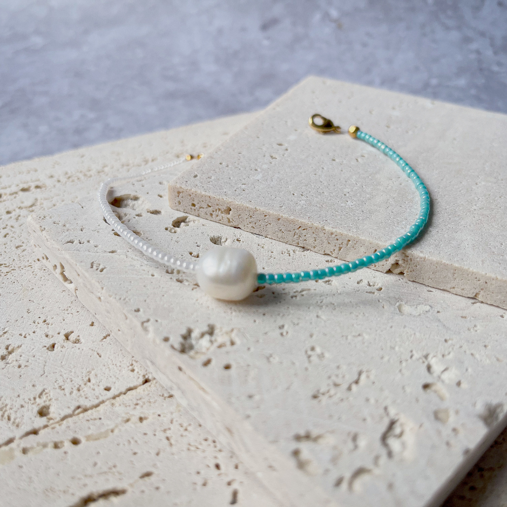 A minimalist bracelet featuring a large single baroque pearl on a string of small white and turquoise glass beads, presented on a corner of two textured aerated concrete blocks. The blocks are placed against a grey background, highlighting the necklace's delicate colors and elegant design. The large pearl serves as a focal point, giving the piece a touch of classic sophistication mixed with a hint of modern color contrast.