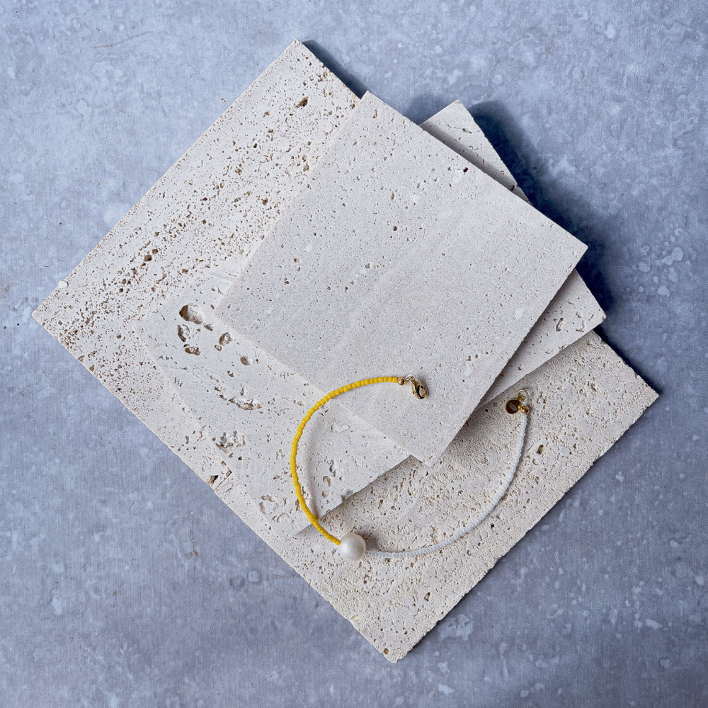 A minimalist bracelet featuring a large single baroque pearl on a string of small white and yellow glass beads, presented on a corner of two textured aerated concrete blocks. The blocks are placed against a grey background, highlighting the necklace's delicate colors and elegant design. The large pearl serves as a focal point, giving the piece a touch of classic sophistication mixed with a hint of modern color contrast.