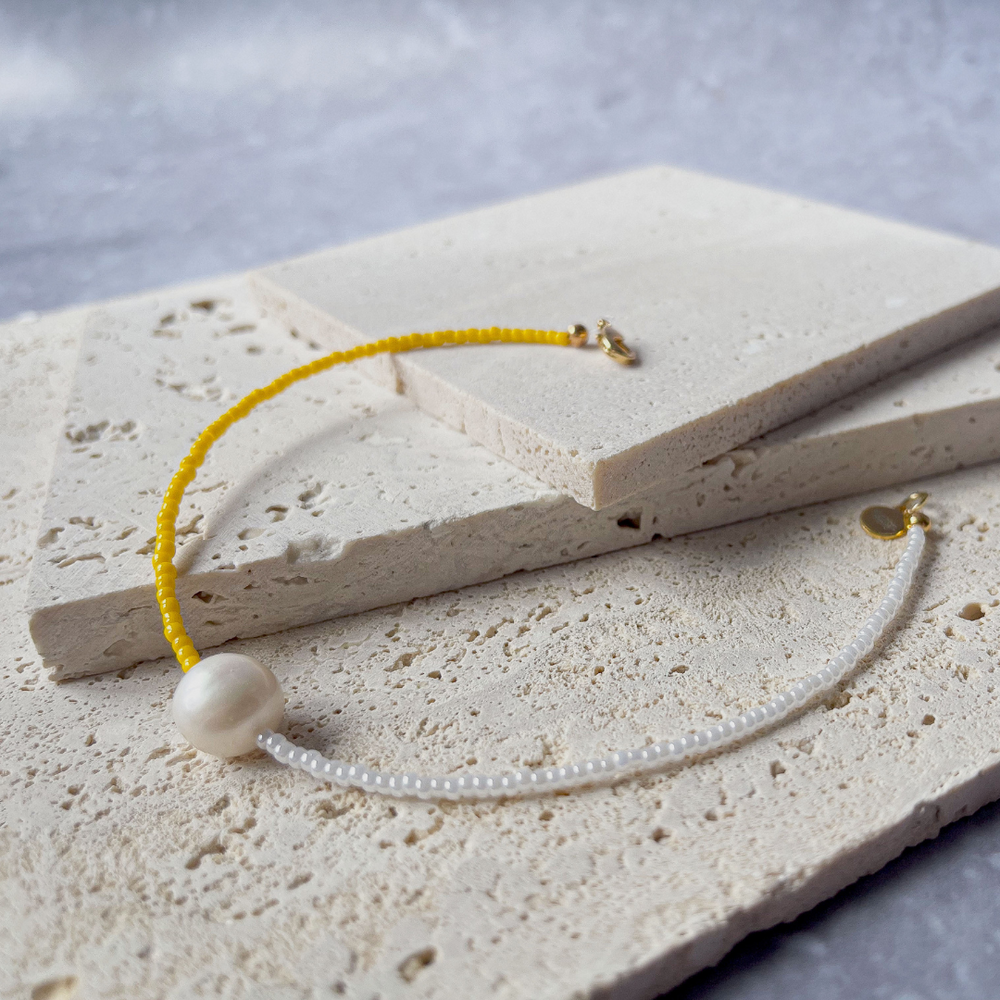 A minimalist bracelet featuring a large single baroque pearl on a string of small white and yellow glass beads, presented on a corner of two textured aerated concrete blocks. The blocks are placed against a grey background, highlighting the necklace's delicate colors and elegant design. The large pearl serves as a focal point, giving the piece a touch of classic sophistication mixed with a hint of modern color contrast.