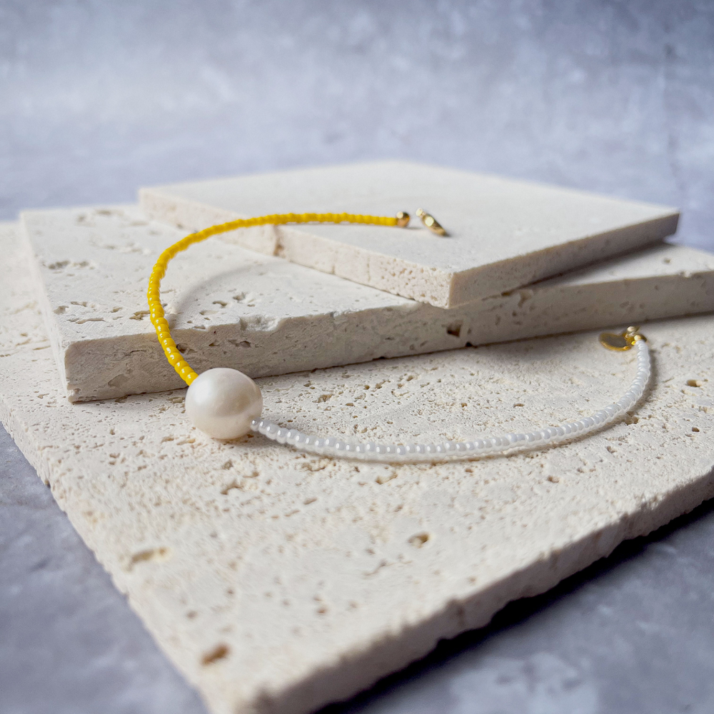 A minimalist bracelet featuring a large single baroque pearl on a string of small white and yellow glass beads, presented on a corner of two textured aerated concrete blocks. The blocks are placed against a grey background, highlighting the necklace's delicate colors and elegant design. The large pearl serves as a focal point, giving the piece a touch of classic sophistication mixed with a hint of modern color contrast.