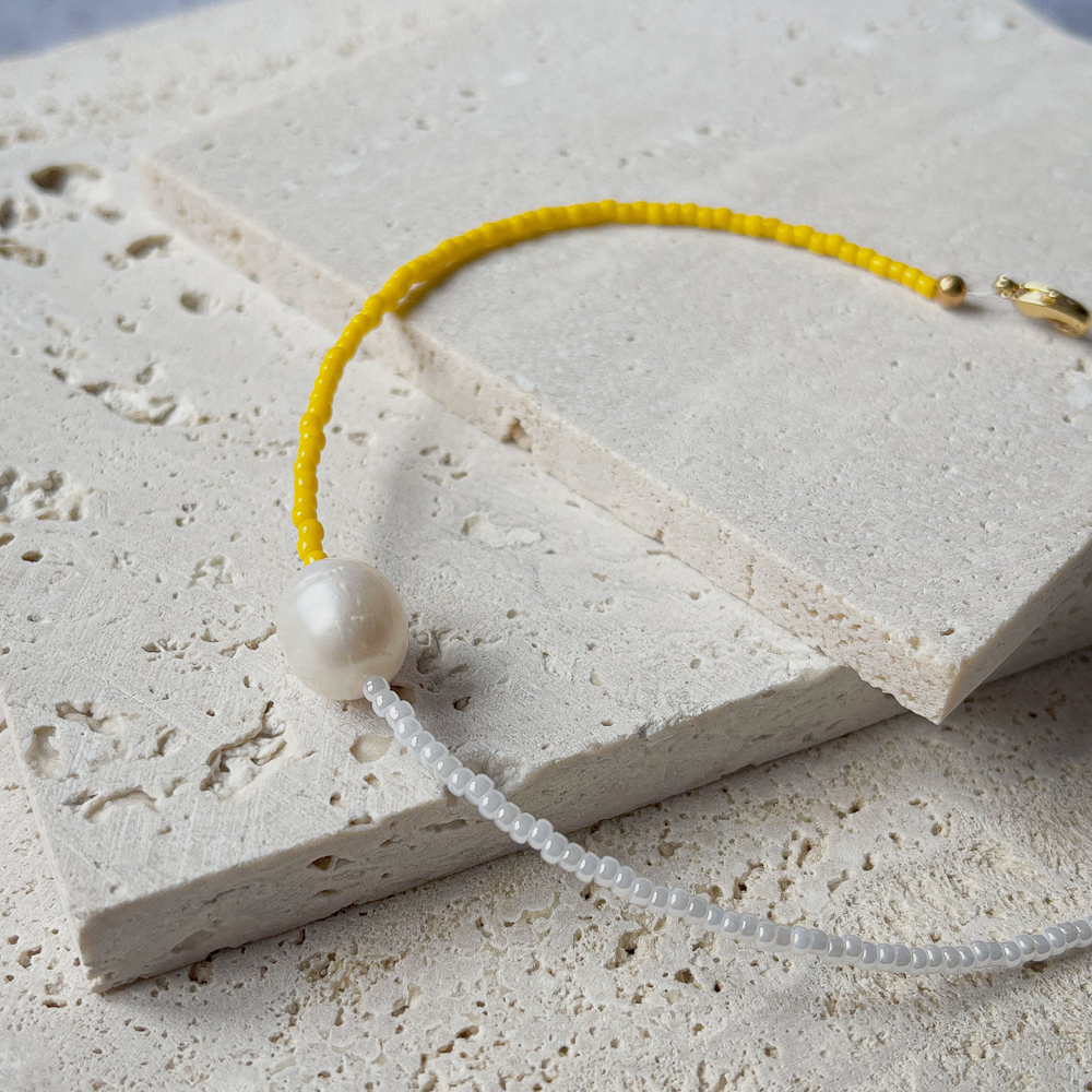 A minimalist bracelet featuring a large single baroque pearl on a string of small white and yellow glass beads, presented on a corner of two textured aerated concrete blocks. The blocks are placed against a grey background, highlighting the necklace's delicate colors and elegant design. The large pearl serves as a focal point, giving the piece a touch of classic sophistication mixed with a hint of modern color contrast.