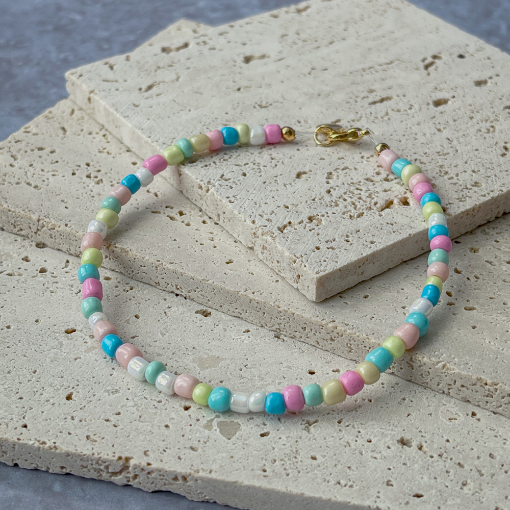 A bracelet with pastel-colored beads in shades of pink, green, blue, and yellow is elegantly draped over two textured concrete blocks. The soft colors of the beads give the piece a playful and gentle appearance, while the gold-tone clasp adds a touch of sophistication. The arrangement on the neutral-toned aerated concrete blocks emphasizes the necklace's colorful charm and suggests a casual yet chic accessory. The background's grey hues contrast with the beads, highlighting their gentle pastel palette.