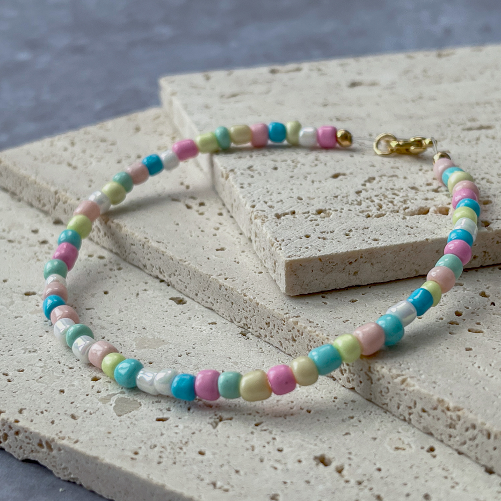 A bracelet with pastel-colored beads in shades of pink, green, blue, and yellow is elegantly draped over two textured concrete blocks. The soft colors of the beads give the piece a playful and gentle appearance, while the gold-tone clasp adds a touch of sophistication. The arrangement on the neutral-toned aerated concrete blocks emphasizes the necklace's colorful charm and suggests a casual yet chic accessory. The background's grey hues contrast with the beads, highlighting their gentle pastel palette.