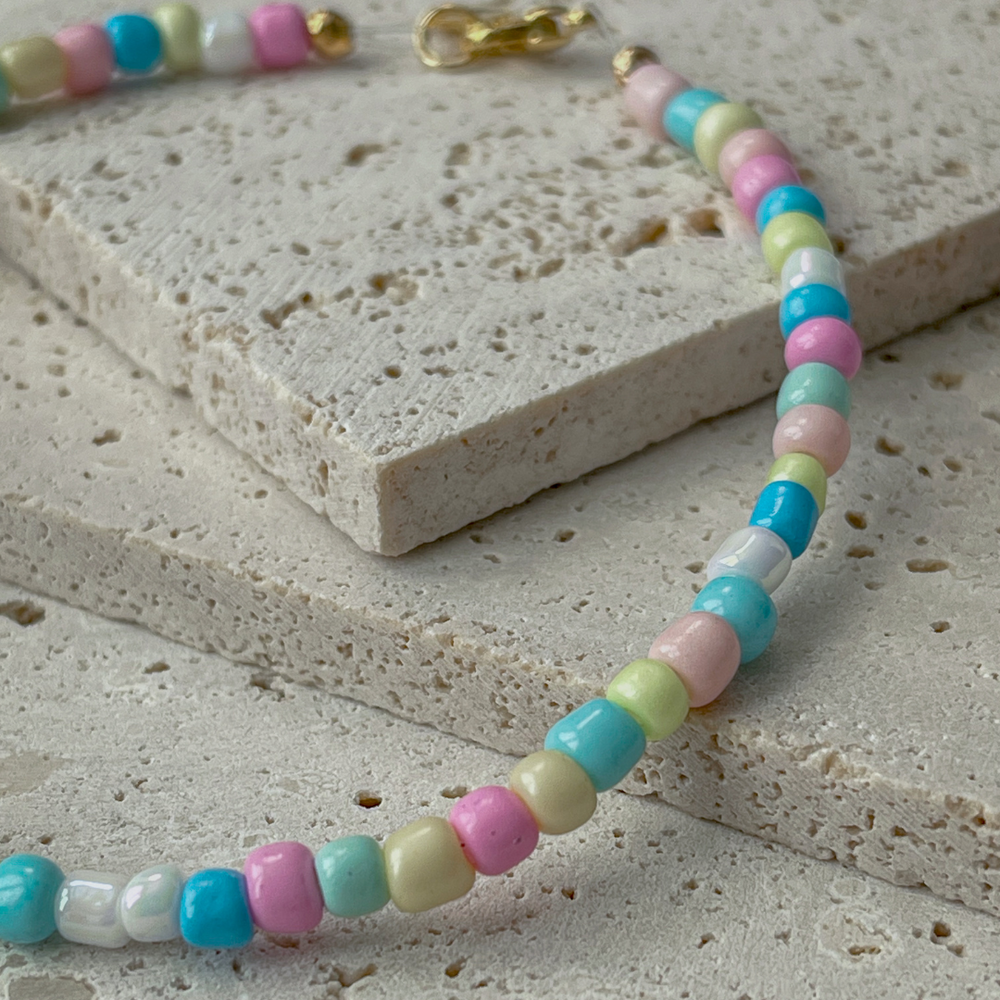 A bracelet with pastel-colored beads in shades of pink, green, blue, and yellow is elegantly draped over two textured concrete blocks. The soft colors of the beads give the piece a playful and gentle appearance, while the gold-tone clasp adds a touch of sophistication. The arrangement on the neutral-toned aerated concrete blocks emphasizes the necklace's colorful charm and suggests a casual yet chic accessory. The background's grey hues contrast with the beads, highlighting their gentle pastel palette.