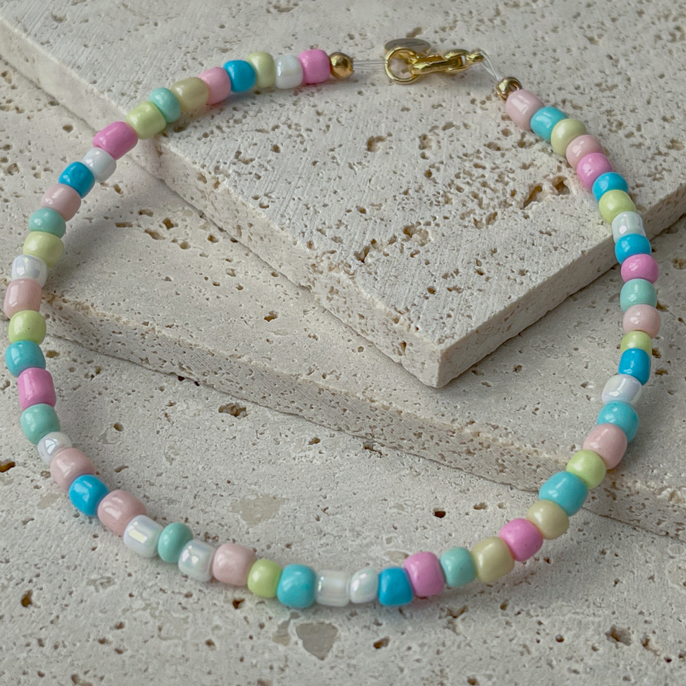 A bracelet with pastel-colored beads in shades of pink, green, blue, and yellow is elegantly draped over two textured concrete blocks. The soft colors of the beads give the piece a playful and gentle appearance, while the gold-tone clasp adds a touch of sophistication. The arrangement on the neutral-toned aerated concrete blocks emphasizes the necklace's colorful charm and suggests a casual yet chic accessory. The background's grey hues contrast with the beads, highlighting their gentle pastel palette.