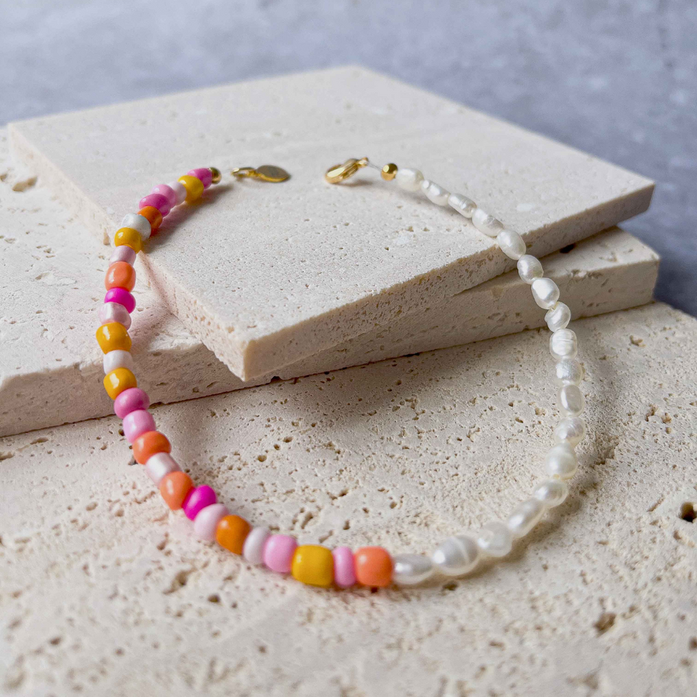 A handmade beaded bracelet with a combination of colorful beads and small white baroque pearls, elegantly placed on two pieces of aerated concrete against a grey textured background. The necklace features a section of vibrant pink, orange, and yellow beads transitioning to translucent white pearls, culminating in a gold-tone clasp with a small brand charm. The overall image showcases the necklace's playful yet sophisticated design.