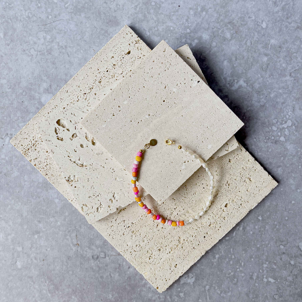 A handmade beaded bracelet with a combination of colorful beads and small white baroque pearls, elegantly placed on two pieces of aerated concrete against a grey textured background. The necklace features a section of vibrant pink, orange, and yellow beads transitioning to translucent white pearls, culminating in a gold-tone clasp with a small brand charm. The overall image showcases the necklace's playful yet sophisticated design.