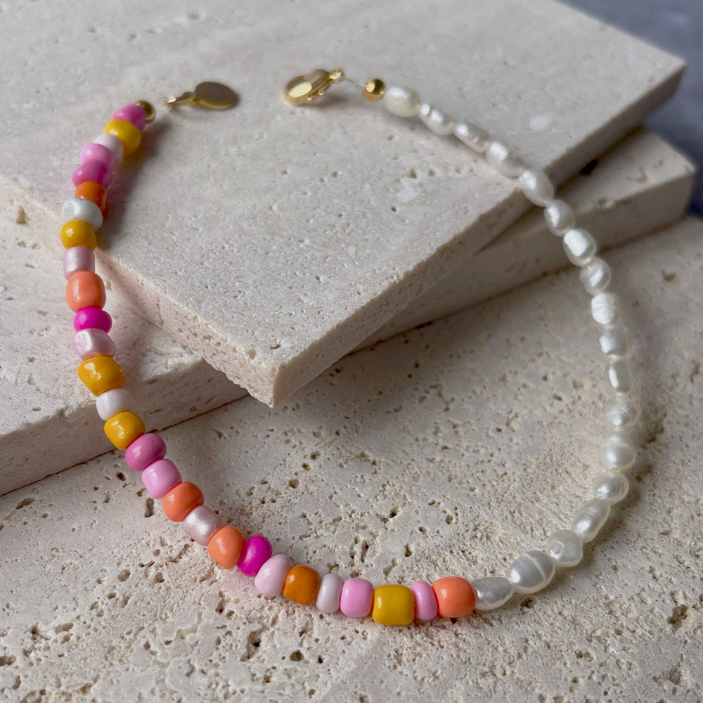 A handmade beaded bracelet with a combination of colorful beads and small white baroque pearls, elegantly placed on two pieces of aerated concrete against a grey textured background. The necklace features a section of vibrant pink, orange, and yellow beads transitioning to translucent white pearls, culminating in a gold-tone clasp with a small brand charm. The overall image showcases the necklace's playful yet sophisticated design.