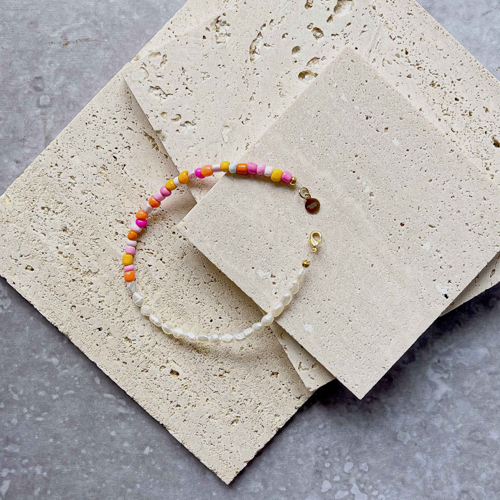 A handmade beaded bracelet with a combination of colorful beads and small white baroque pearls, elegantly placed on two pieces of aerated concrete against a grey textured background. The necklace features a section of vibrant pink, orange, and yellow beads transitioning to translucent white pearls, culminating in a gold-tone clasp with a small brand charm. The overall image showcases the necklace's playful yet sophisticated design.