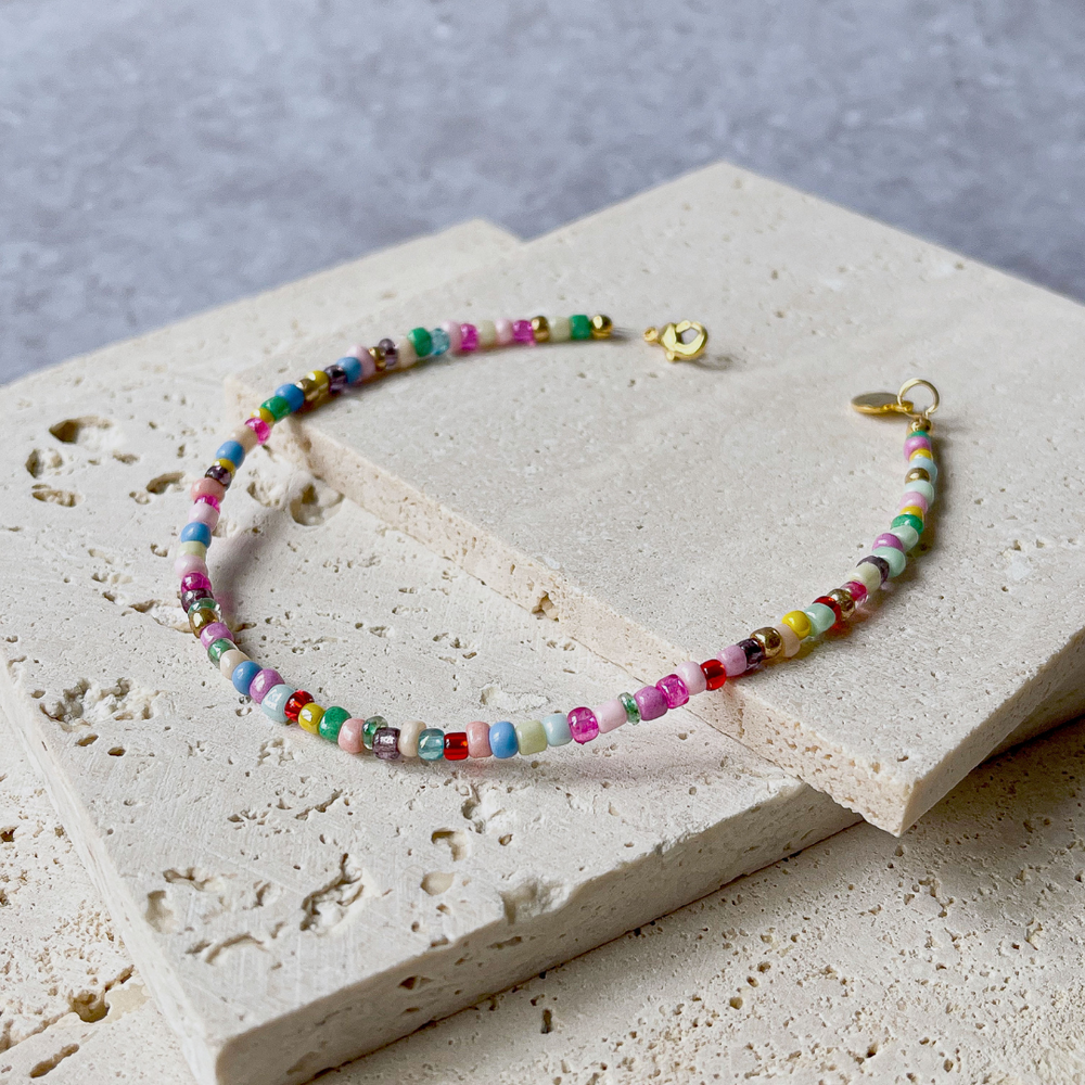 A colorful beaded bracelet with a vibrant array of multicolored beads, displayed across the corner of a textured aerated concrete block. The necklace showcases a playful mix of red, green, blue, yellow, and pink beads with interspersed white ones, creating a rainbow effect. A gold-tone clasp and charm add a touch of elegance to the piece. The bracelet is set against a grey background that accentuates its bright colors and joyful design.