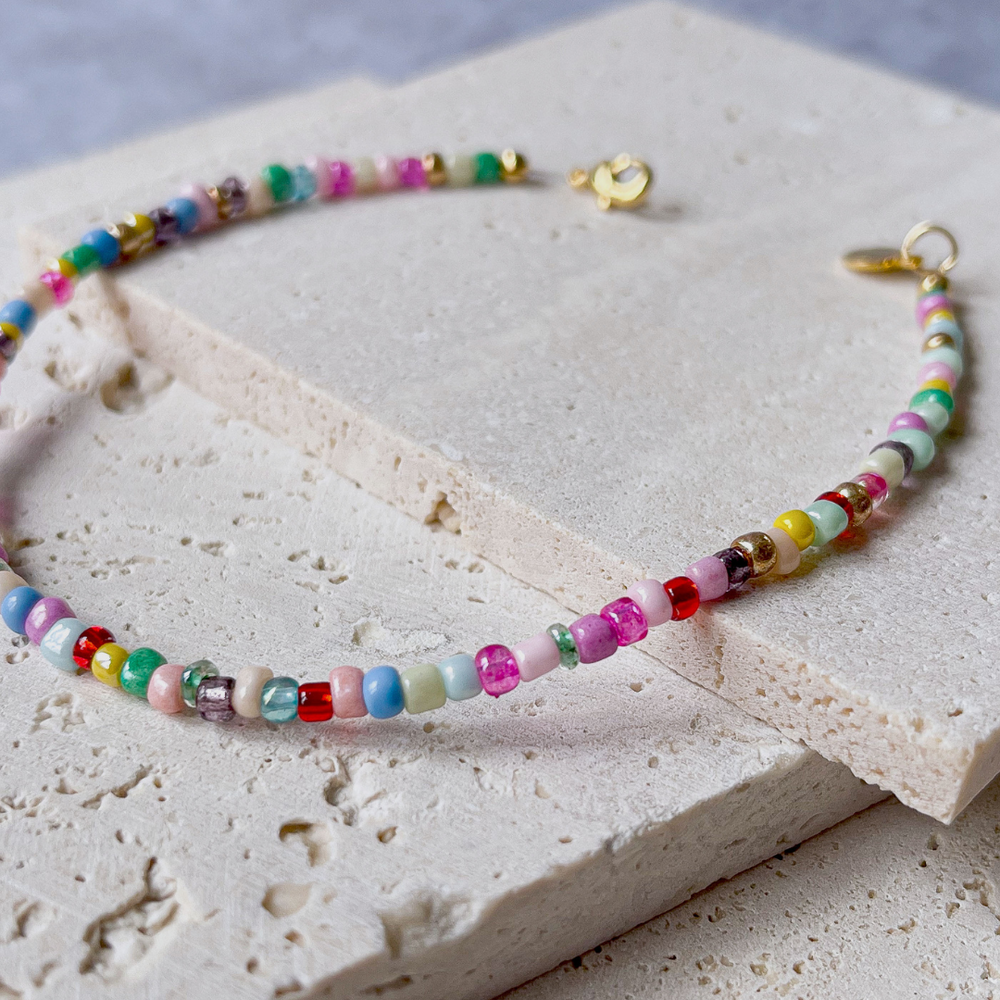 A colorful beaded bracelet with a vibrant array of multicolored beads, displayed across the corner of a textured aerated concrete block. The necklace showcases a playful mix of red, green, blue, yellow, and pink beads with interspersed white ones, creating a rainbow effect. A gold-tone clasp and charm add a touch of elegance to the piece. The bracelet is set against a grey background that accentuates its bright colors and joyful design.