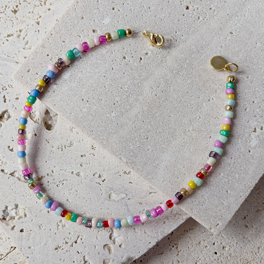 A colorful beaded bracelet with a vibrant array of multicolored beads, displayed across the corner of a textured aerated concrete block. The necklace showcases a playful mix of red, green, blue, yellow, and pink beads with interspersed white ones, creating a rainbow effect. A gold-tone clasp and charm add a touch of elegance to the piece. The bracelet is set against a grey background that accentuates its bright colors and joyful design.