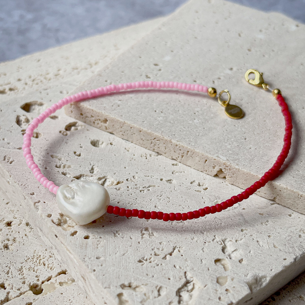 A minimalist bracelet featuring a large single baroque pearl on a string of small red and bright pink glass beads, presented on a corner of two textured aerated concrete blocks. The blocks are placed against a grey background, highlighting the necklace's delicate colors and elegant design. The large pearl serves as a focal point, giving the piece a touch of classic sophistication mixed with a hint of modern color contrast.