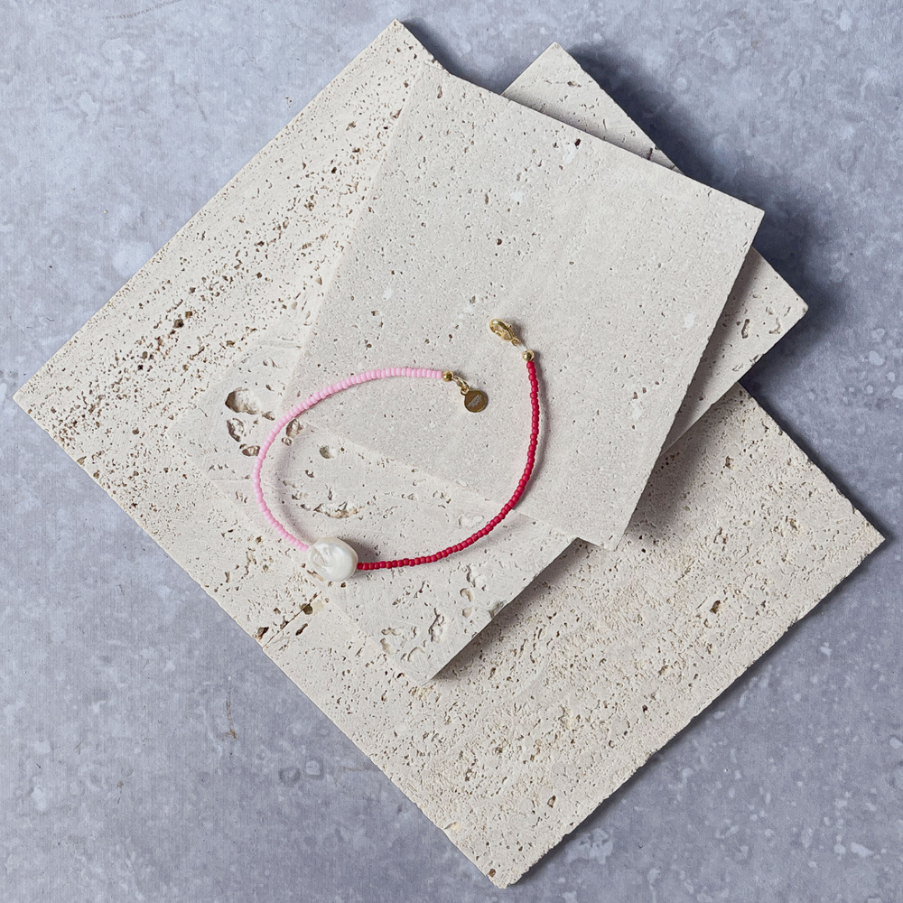 A minimalist bracelet featuring a large single baroque pearl on a string of small red and bright pink glass beads, presented on a corner of two textured aerated concrete blocks. The blocks are placed against a grey background, highlighting the necklace's delicate colors and elegant design. The large pearl serves as a focal point, giving the piece a touch of classic sophistication mixed with a hint of modern color contrast.