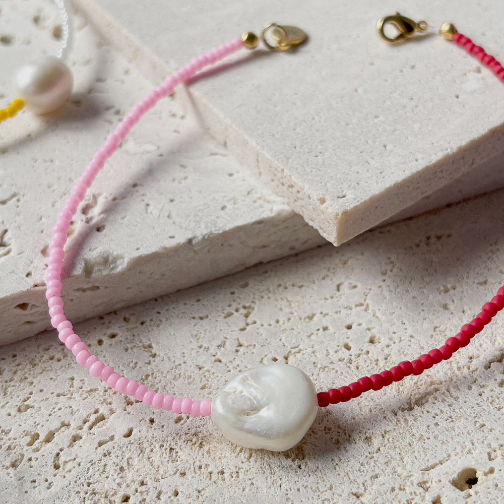 A minimalist bracelet featuring a large single baroque pearl on a string of small red and bright pink glass beads, presented on a corner of two textured aerated concrete blocks. The blocks are placed against a grey background, highlighting the necklace's delicate colors and elegant design. The large pearl serves as a focal point, giving the piece a touch of classic sophistication mixed with a hint of modern color contrast.