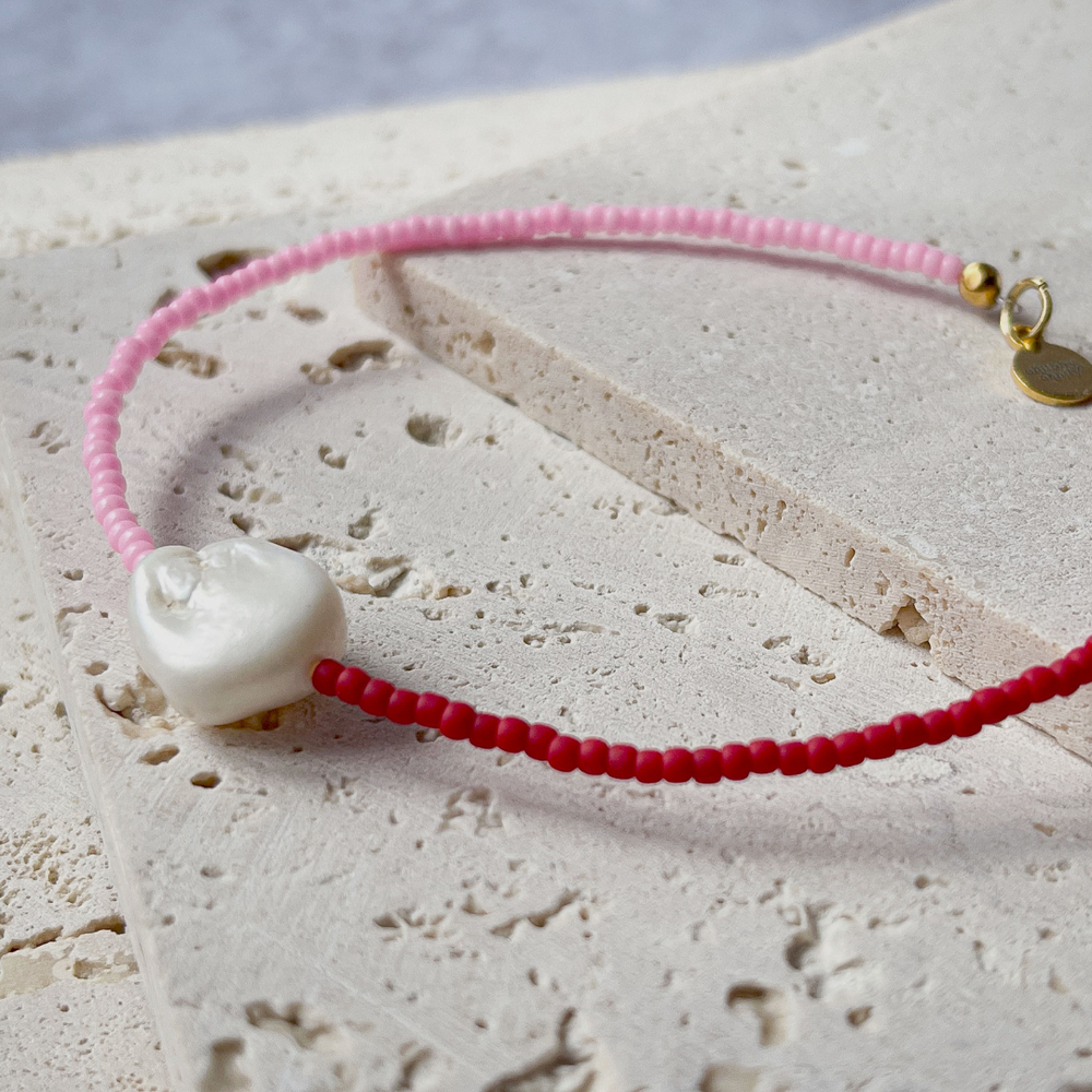 A minimalist bracelet featuring a large single baroque pearl on a string of small red and bright pink glass beads, presented on a corner of two textured aerated concrete blocks. The blocks are placed against a grey background, highlighting the necklace's delicate colors and elegant design. The large pearl serves as a focal point, giving the piece a touch of classic sophistication mixed with a hint of modern color contrast.