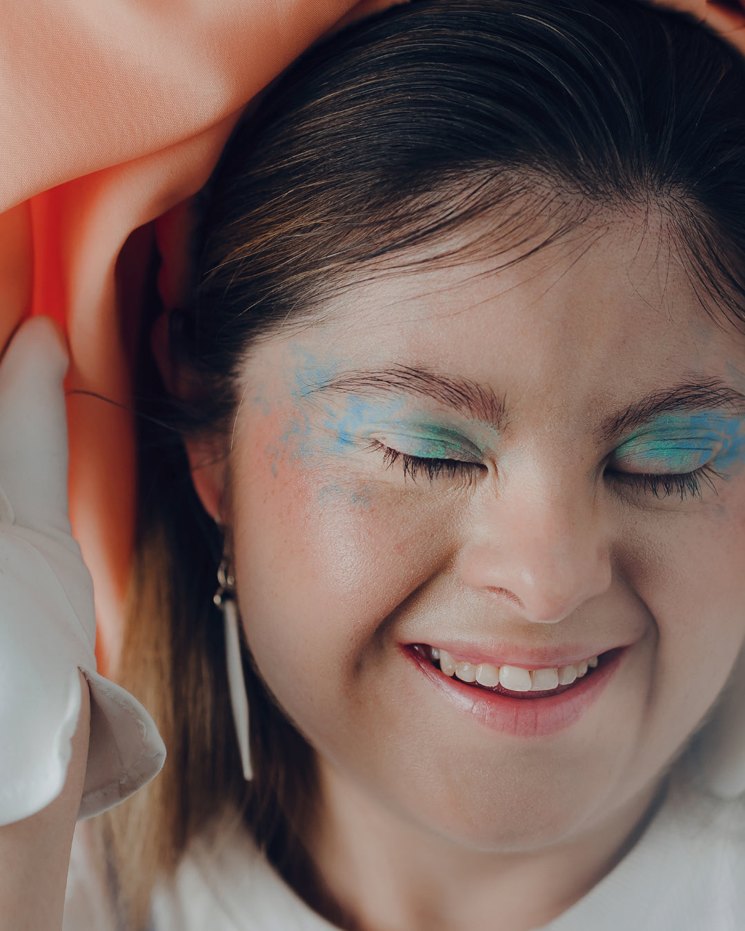 A close-up portrait of a beautiful woman with Down Syndrome. She has her eyes closed, wears bright blue eyeshadow and smiles.