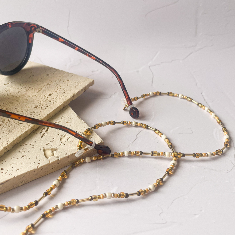 A pair of tortoiseshell sunglasses with round frames is partially resting on two beige aerated concrete blocks. A delicate eyeglass chain with white and golden beads loops around one of the blocks, creating a heart shape on a white marbled surface. The image conveys a fashionable and practical accessory for eyewear, with a focus on handmade craftsmanship.