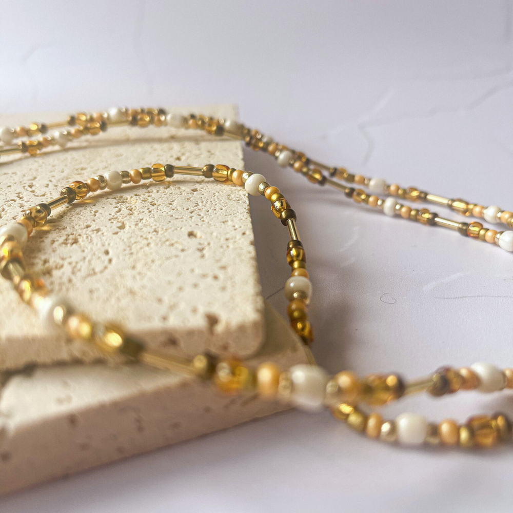 A pair of tortoiseshell sunglasses with round frames is partially resting on two beige aerated concrete blocks. A delicate eyeglass chain with white and golden beads loops around one of the blocks, creating a heart shape on a white marbled surface. The image conveys a fashionable and practical accessory for eyewear, with a focus on handmade craftsmanship.