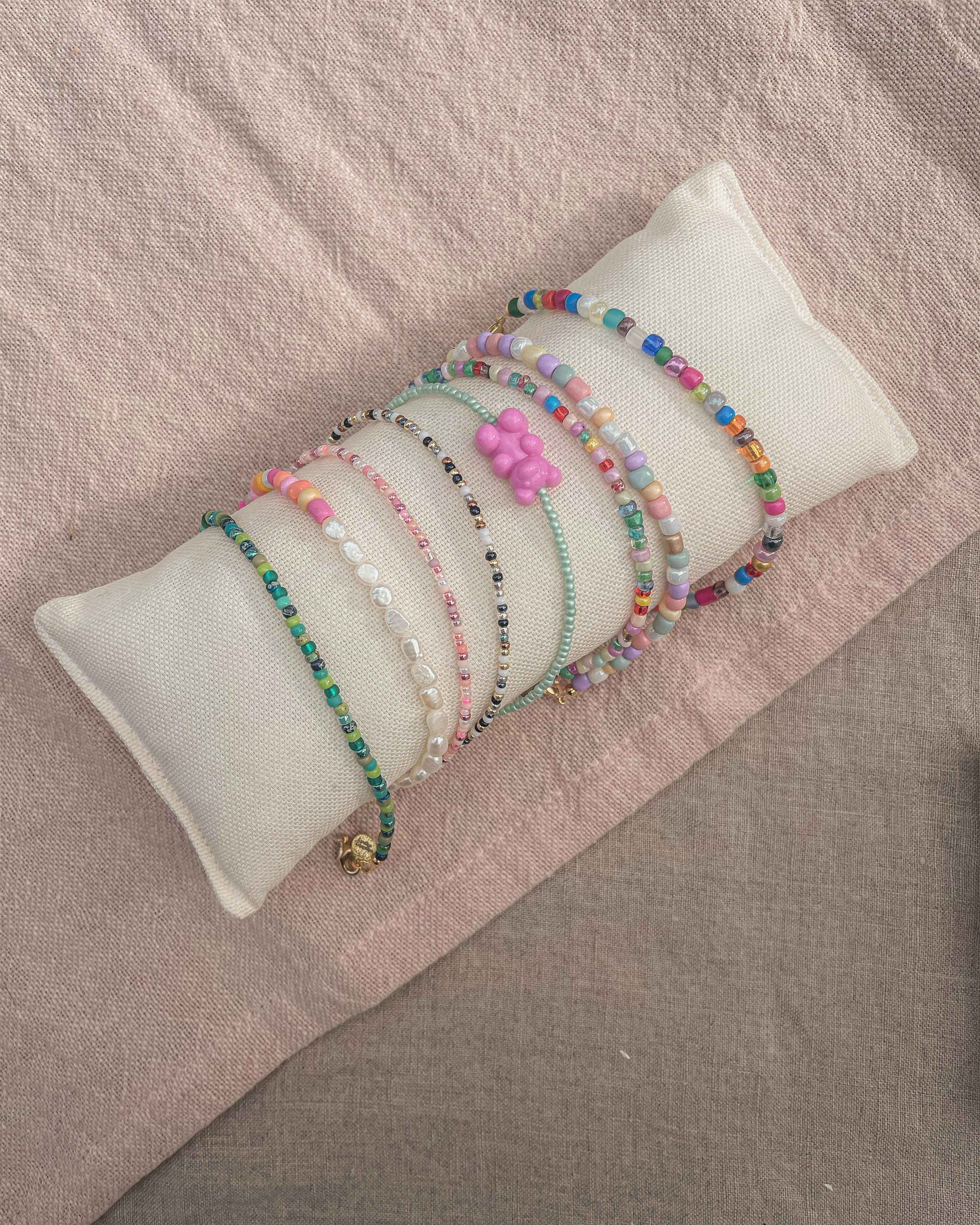 A collection of colorful beaded bracelets displayed on a white cylindrical jewelry stand, which is set against a pinkish-beige fabric background. The bracelets feature a variety of beads including multicolored and patterned designs, translucent and opaque finishes, with one showcasing a prominent pink flower-shaped bead. The arrangement suggests a playful and youthful fashion accessory selection.